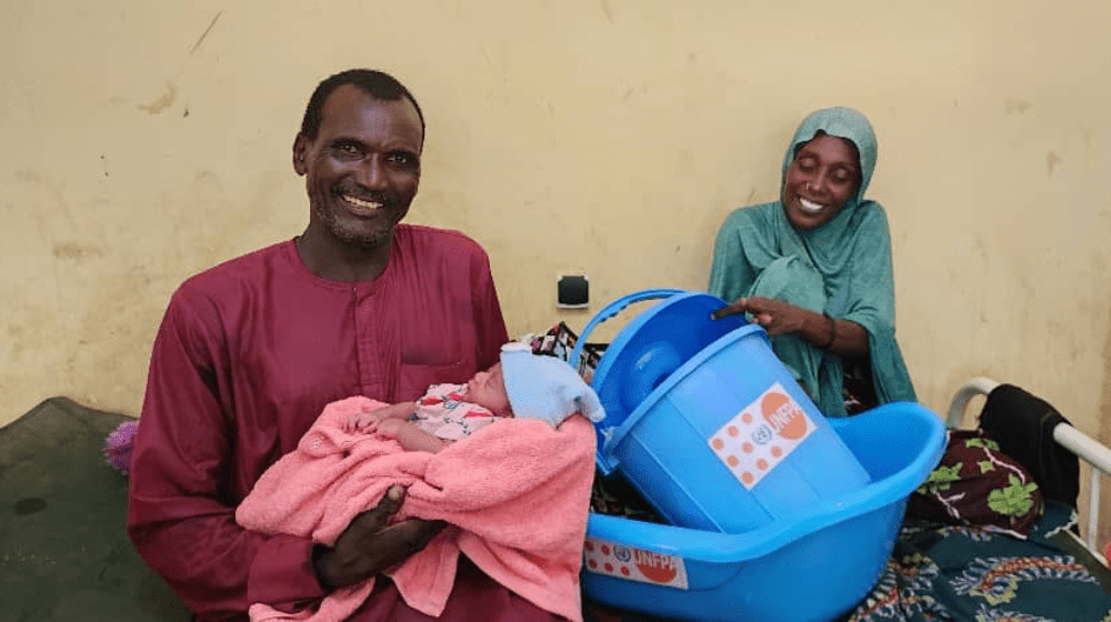 A man wearing red holds a newborn baby. Behind him, seated on a bed, is the baby’s mother, wearing blue, holding a blue plastic tub with a UNFPA logo.